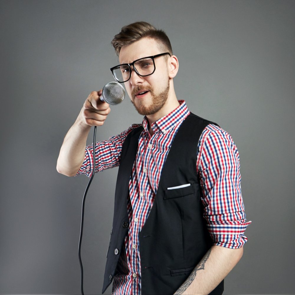 Karaoke man sings the song to microphone, singer with beard on grey background. Funny man in glasses