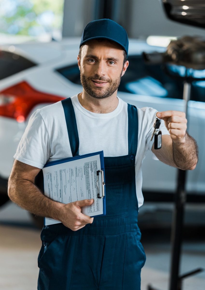 happy car mechanic looking at camera and holding clipboard and key near car