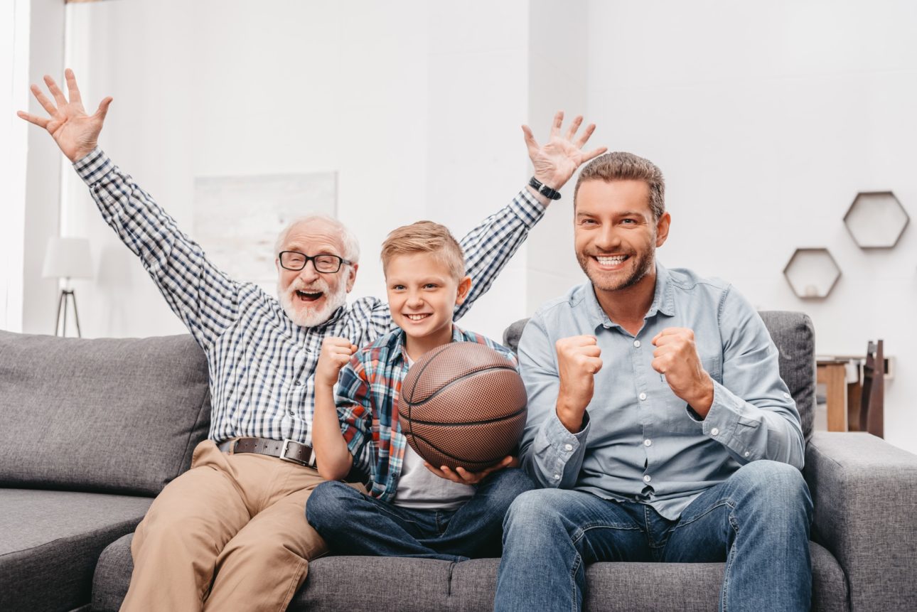 Little boy on couch with grandfather and father, cheering for a basketball game and holding a