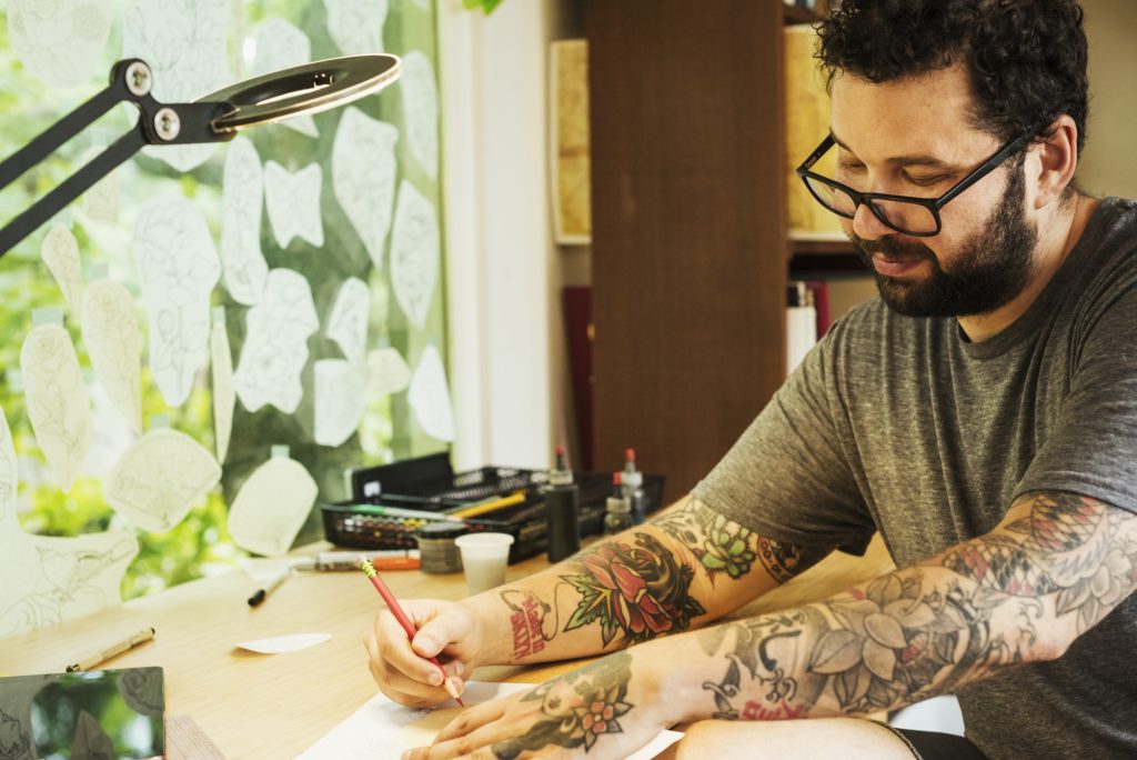 Bearded man with tattoos on his arms, sitting at a desk, drawing.