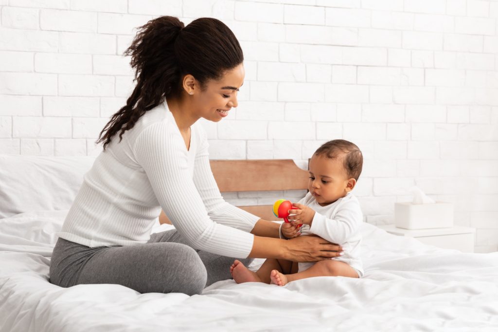 Black Mother Playing With Baby Giving Toy Sitting In Bedroom