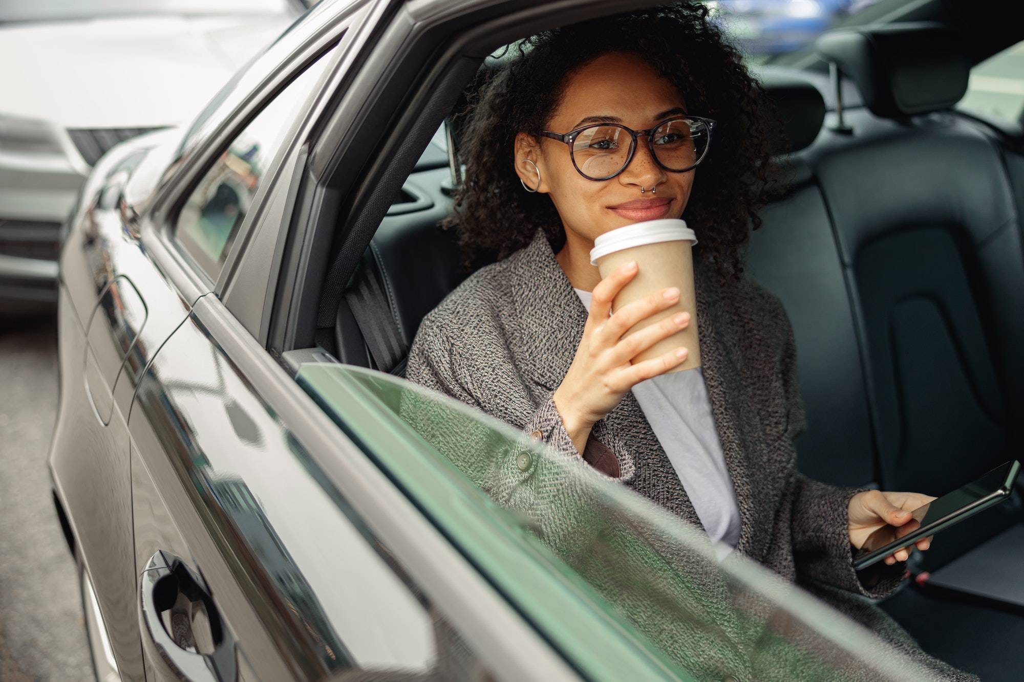Smiling businesswoman with coffee use phone and sitting on backseat of taxi