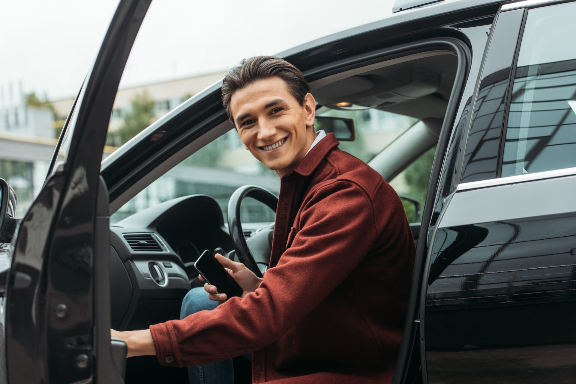 Smiling taxi driver sitting in car with open door and holding smartphone