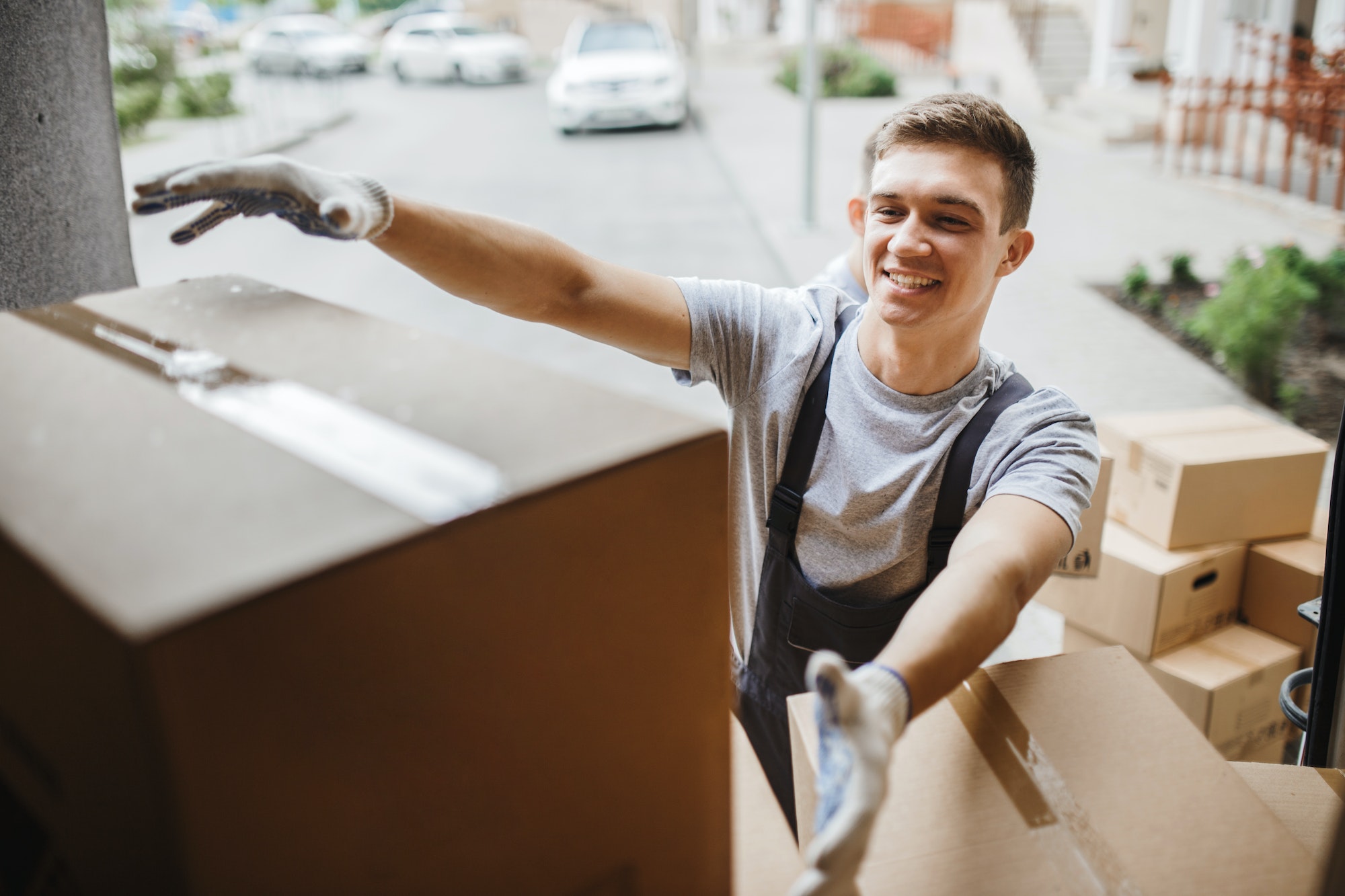 A young handsome smiling mover wearing uniform is reaching for the box while unloading the van full