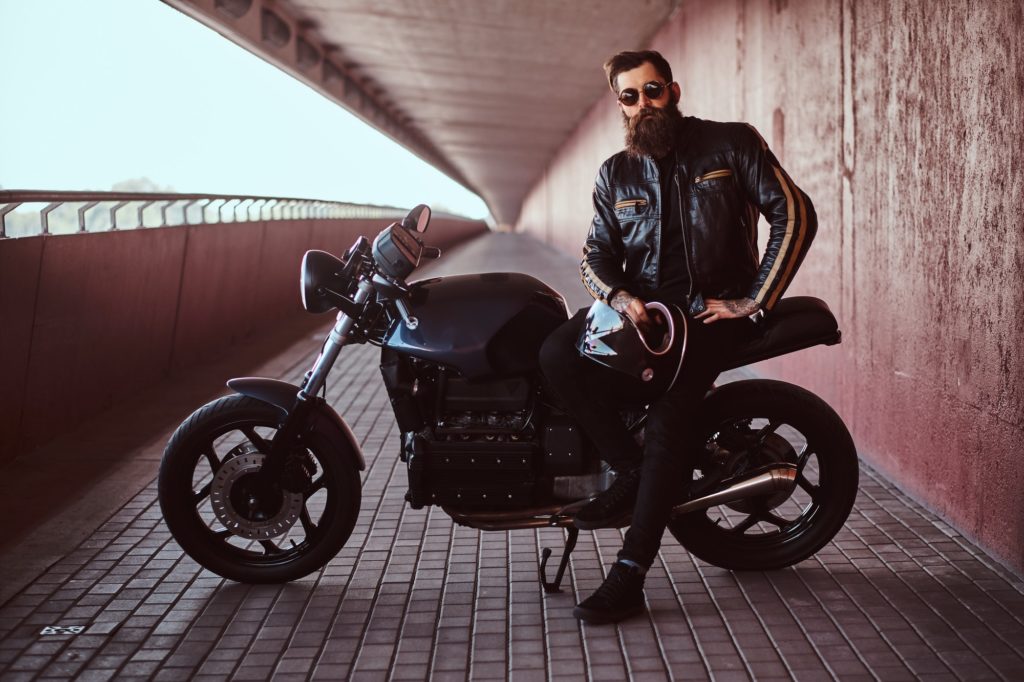 Stylish bearded male dressed in a black leather jacket sitting on his custom-made retro motorcycle