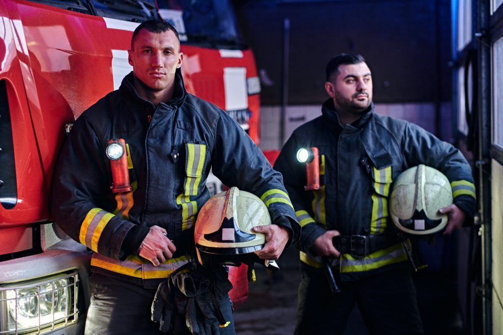Two firemen standing near the fire truck at night in a fire depot