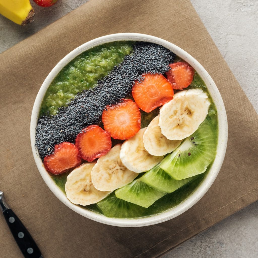 top view of smoothie bowl with fresh fruits, spoon and ingredients on grey background