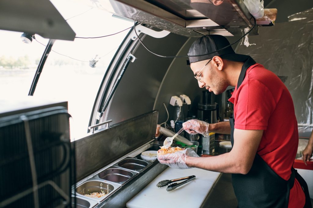 Young man in uniform cooking tasty hotdogs in food truck