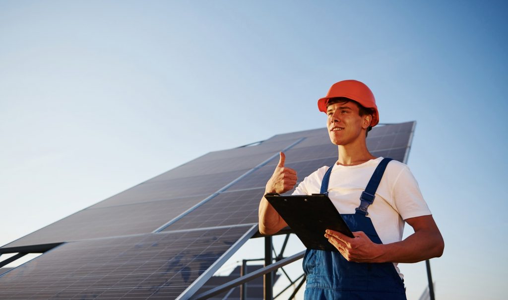 Shows thumb up. Male worker in blue uniform outdoors with solar batteries at sunny day