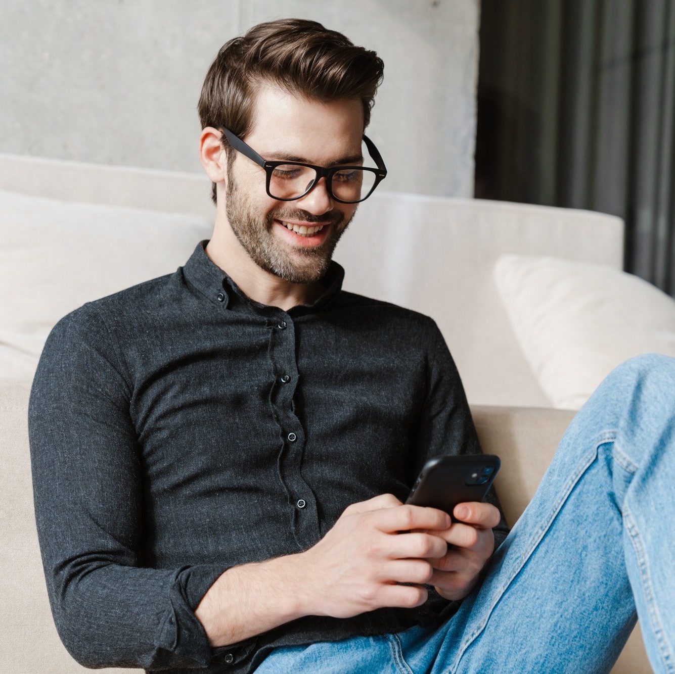 Cheerful unshaven man using smartphone while sitting on floor