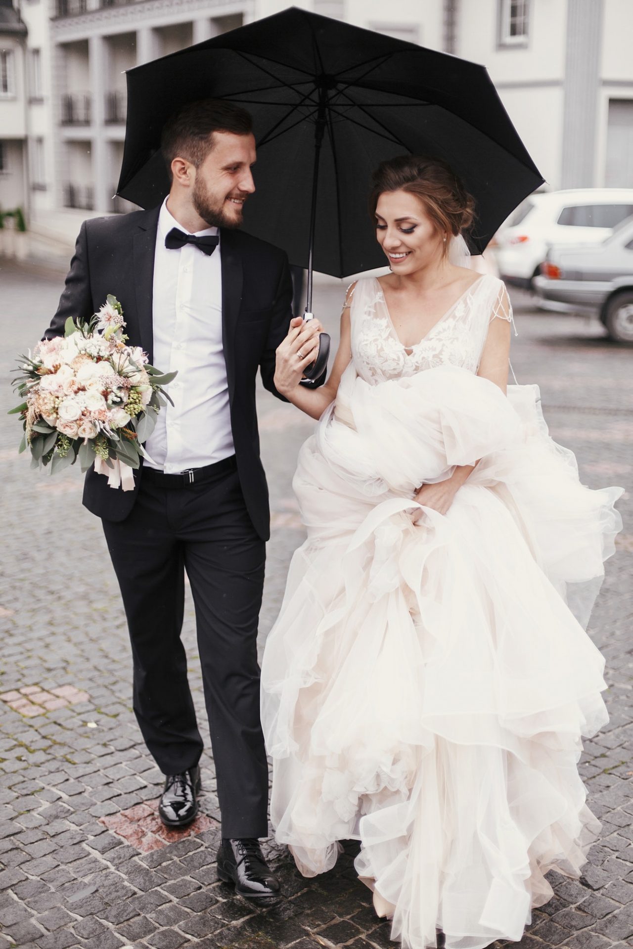 Gorgeous bride and stylish groom walking under umbrella in rainy street and smiling