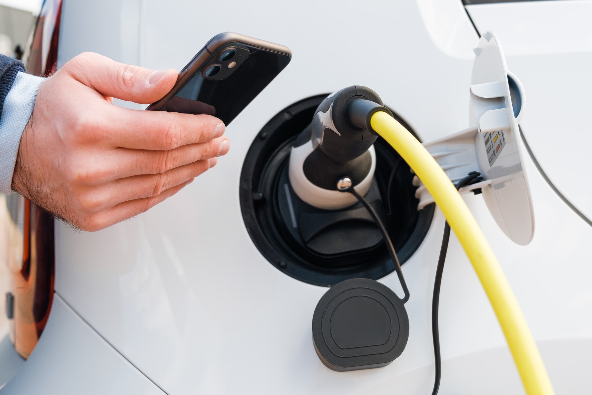 Close up man hand holding a smartphone near charging electric car, EV car