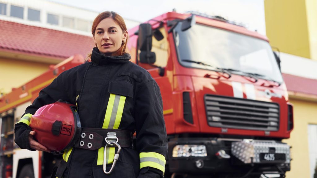 Female firefighter in protective uniform standing near truck