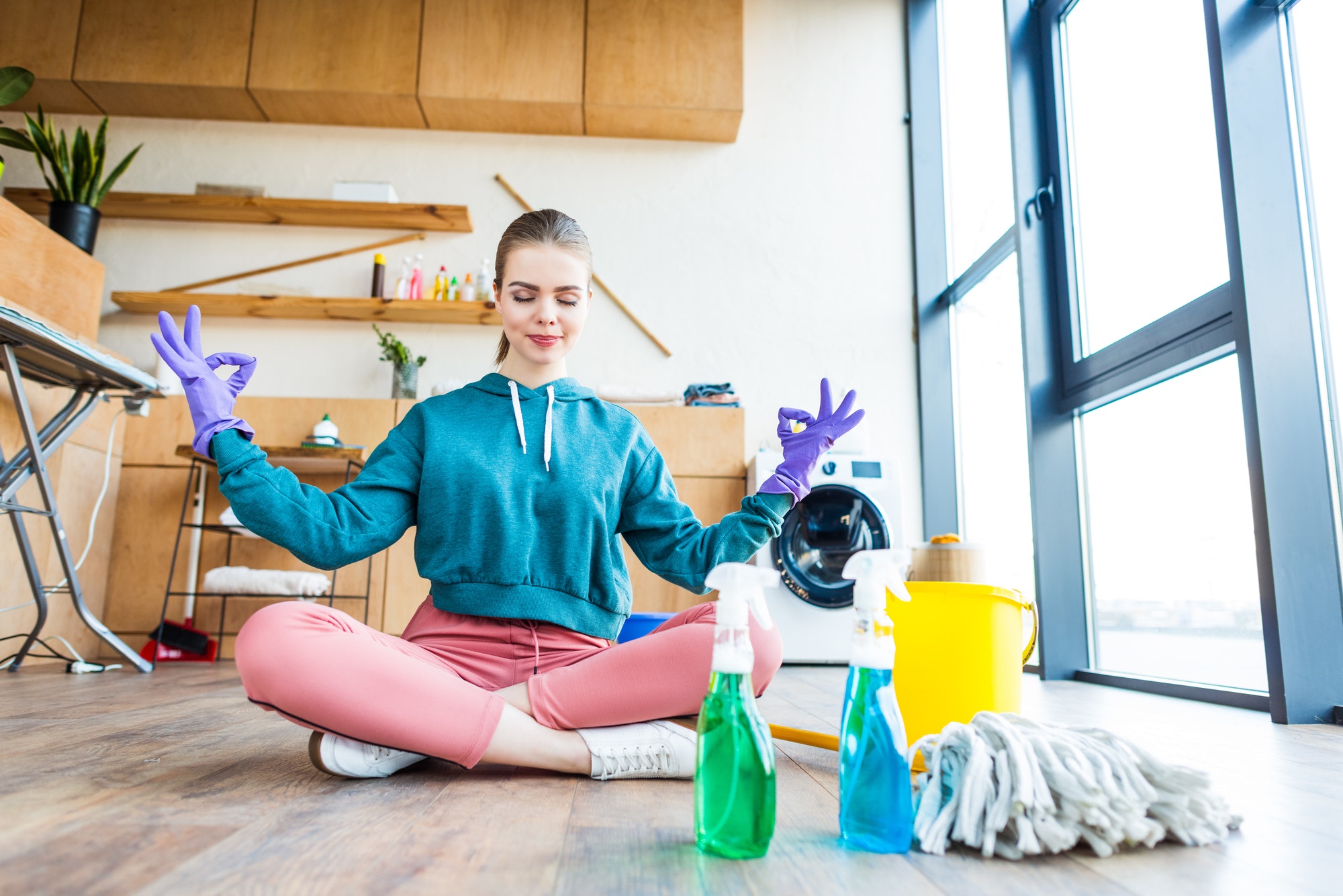 smiling young woman sitting on floor and meditating while cleaning house