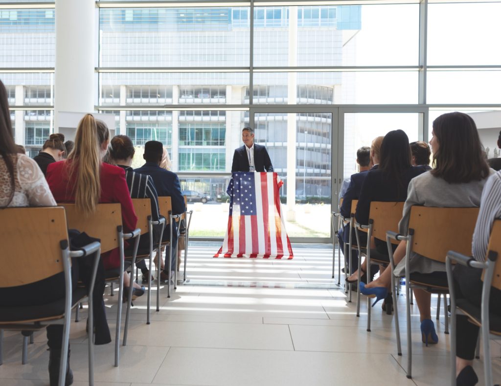 Male speaker speaks in a business seminar in office building with american flag on his mic stand