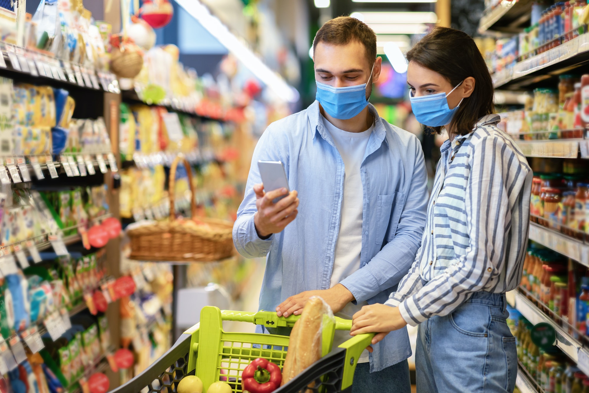 Young couple in masks with phone shopping in supermarket