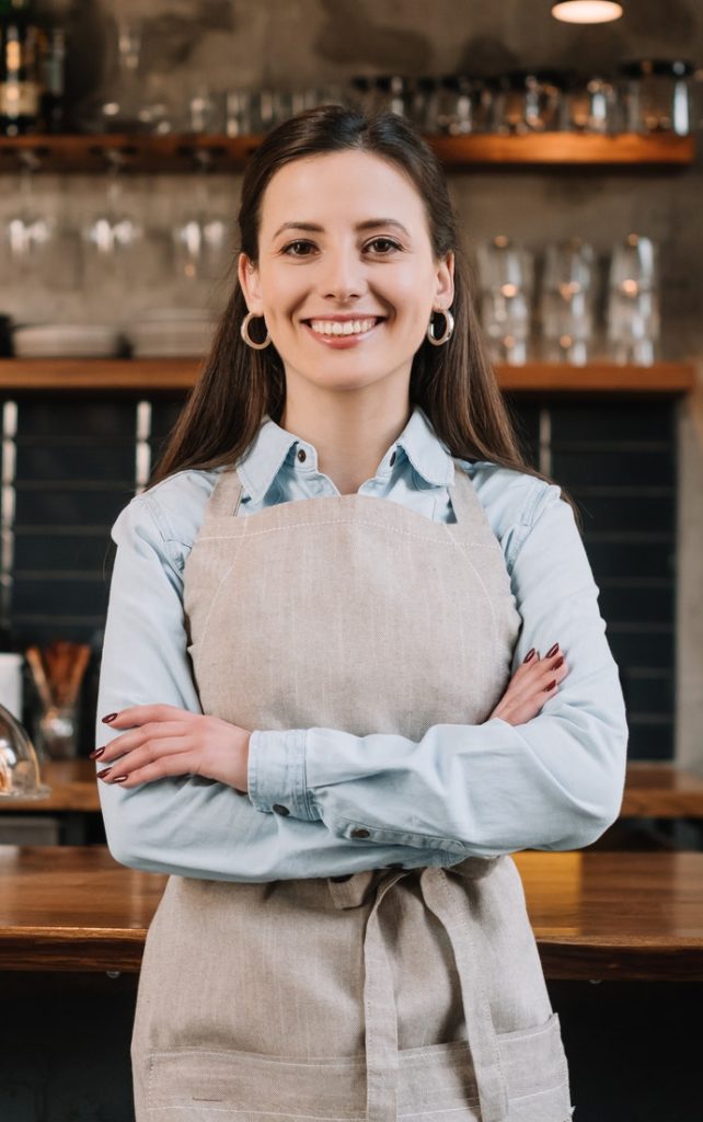 smiling barista in apron standing with crossed arms near bar counter