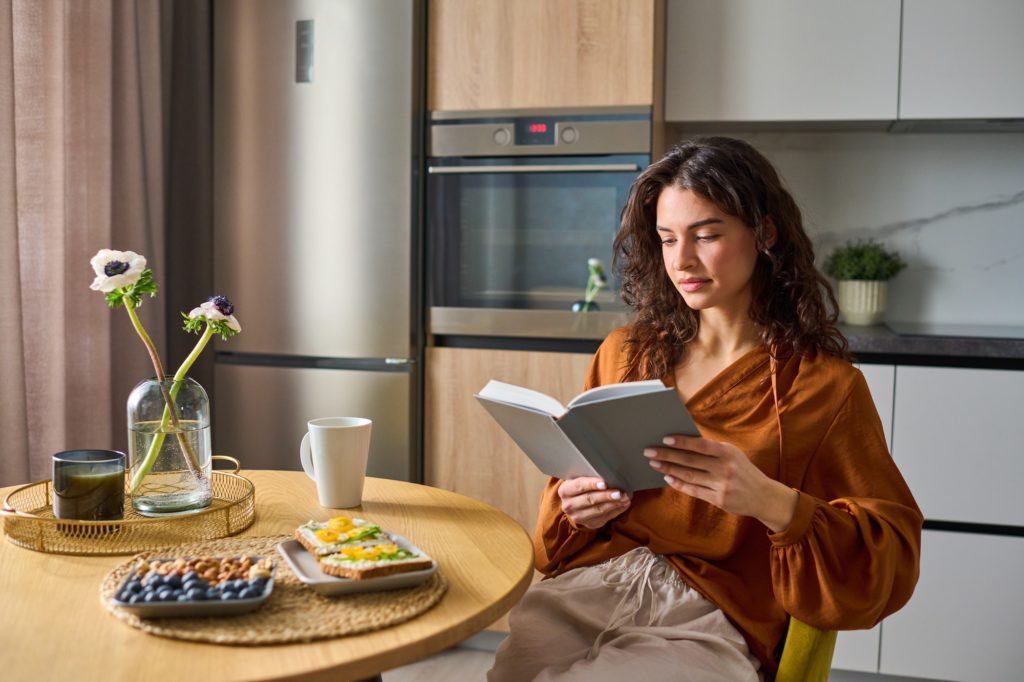 Young relaxed woman in casualwear reading book while sitting by kitchen table