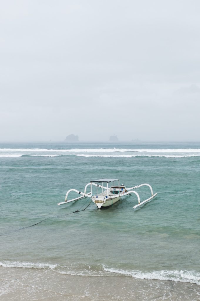 One white traditional Bali fishing boat on big waves in storm and raining. Candidasa beach