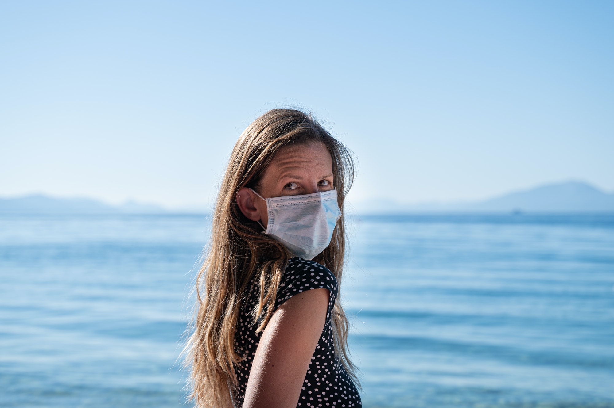 Young woman with medical protective mask standing by the sea.