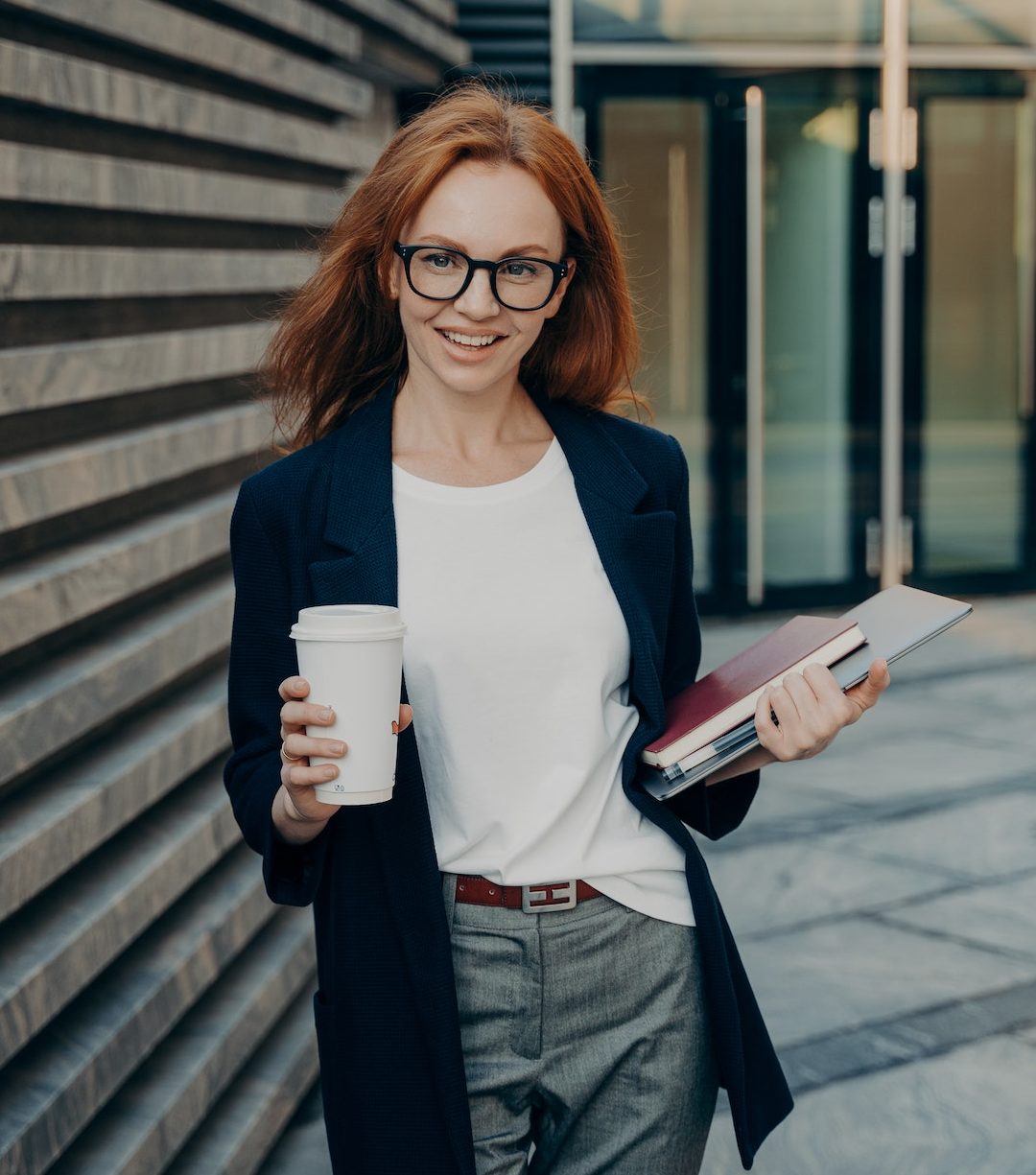 Successful ginger businesswoman standing outdoors near office with laptop, notebook and coffee