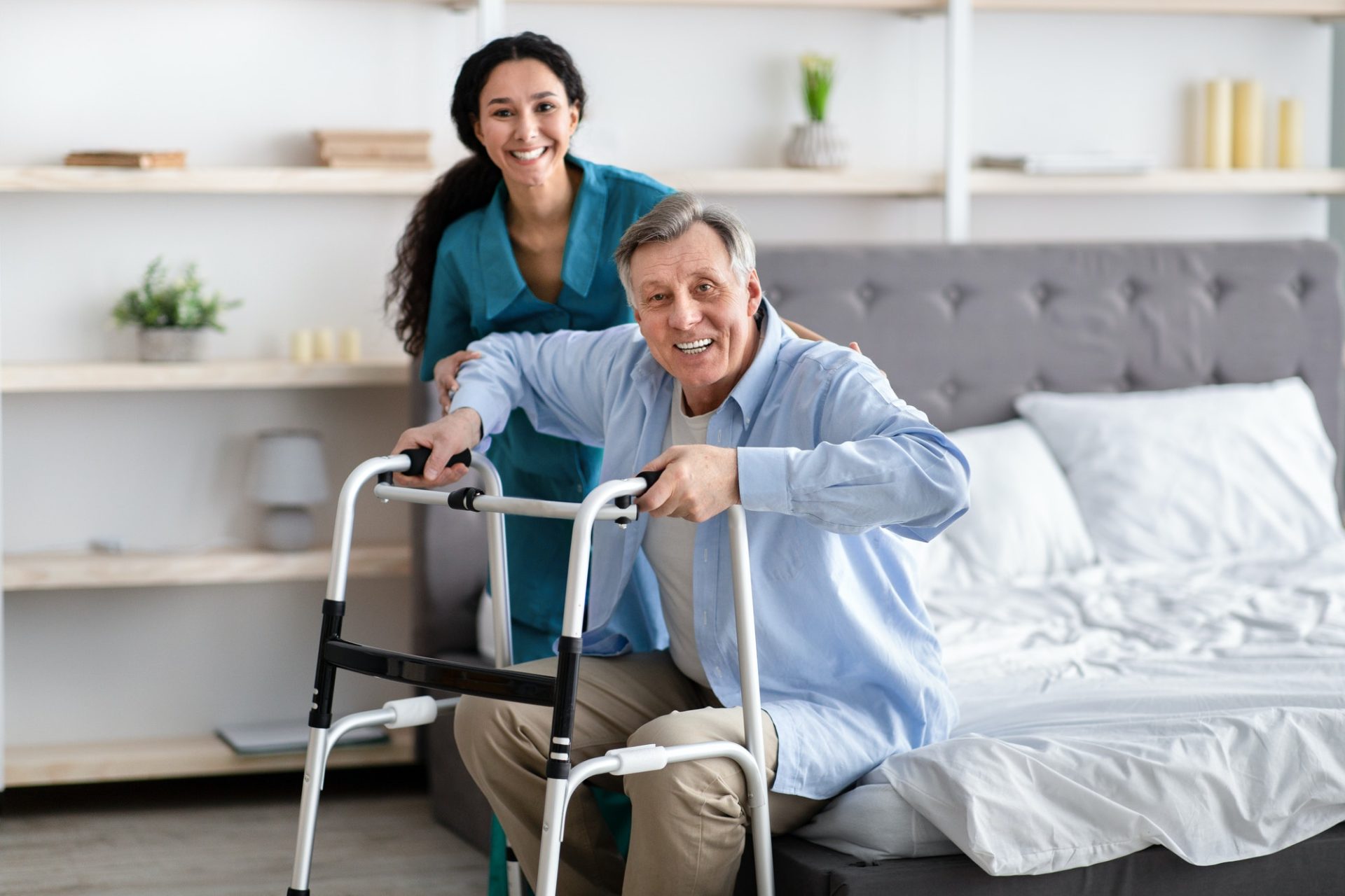 Female nurse helping elderly male with walking frame stand up from bed at home. Professional care