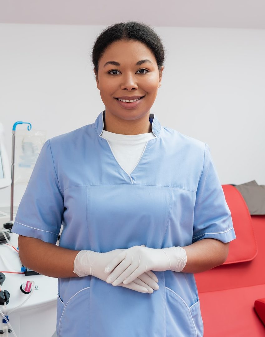 cheerful multiracial nurse in blue uniform and latex gloves looking at camera near automated