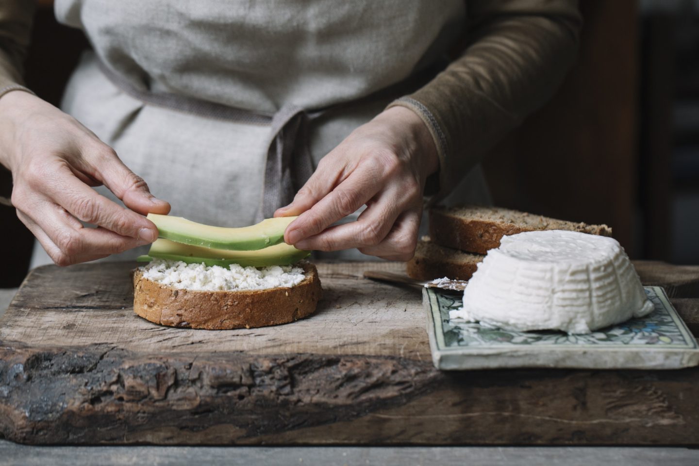 Woman placing slices of avocado onto sliced bread with ricotta, mid section