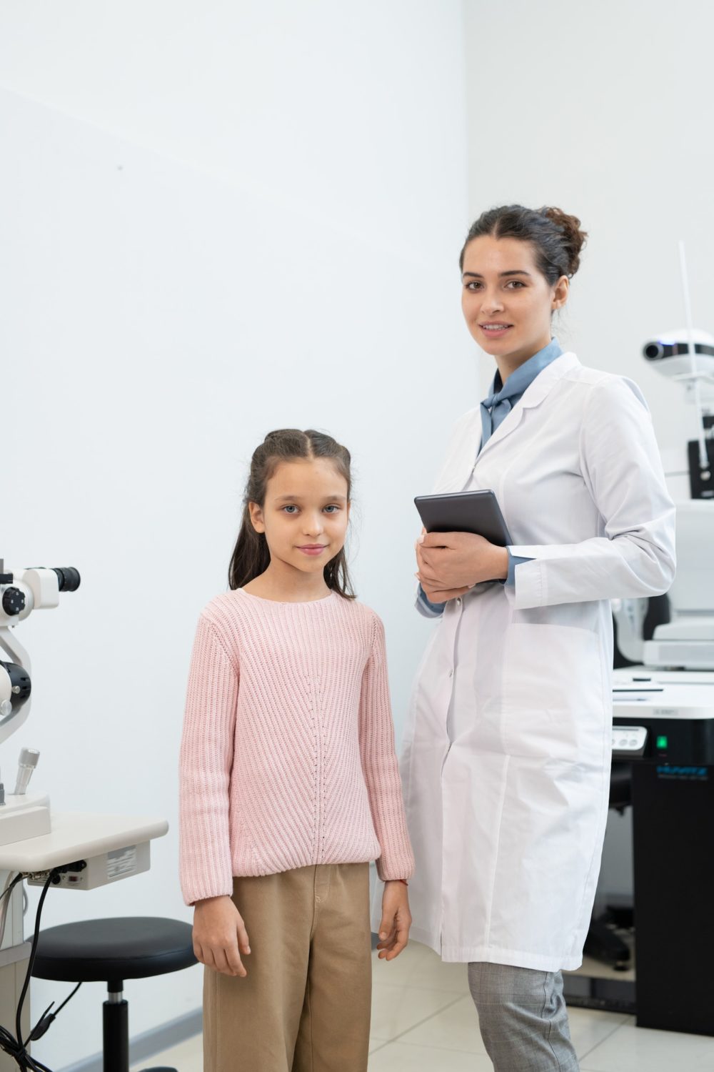 Happy young female ophthalmologist in whitecoat with tablet and schoolgirl
