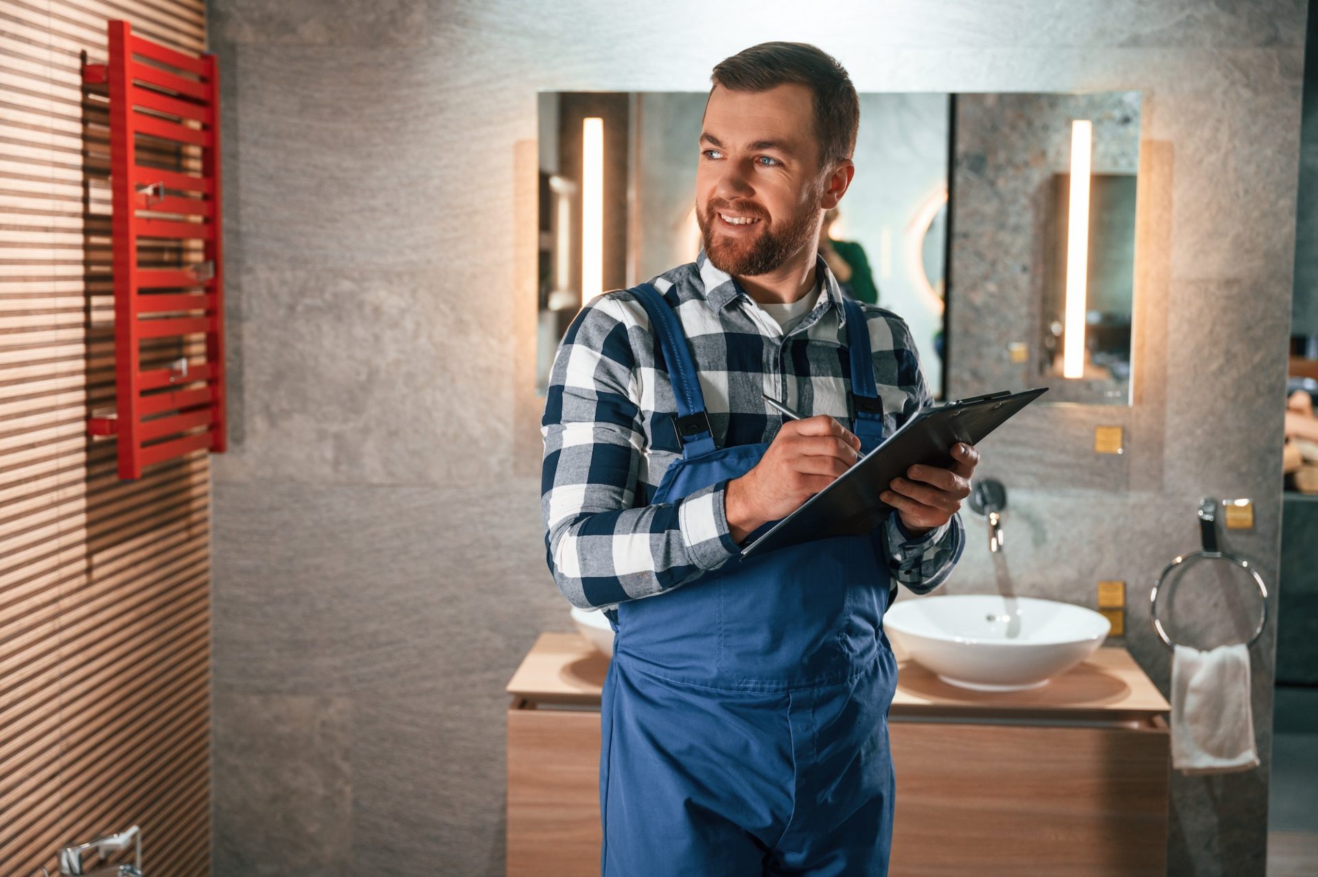 With notepad in hands. Plumber in blue uniform is at work in the bathroom
