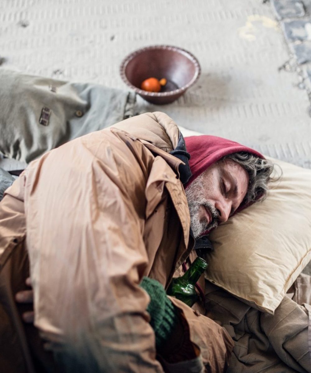 Homeless beggar man lying on the ground outdoors in city, sleeping.
