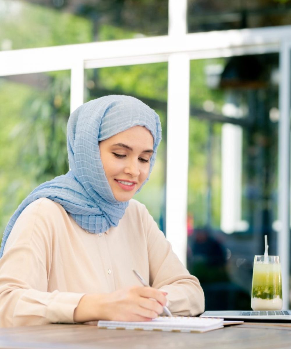 Young muslim student in hijab making working notes by table
