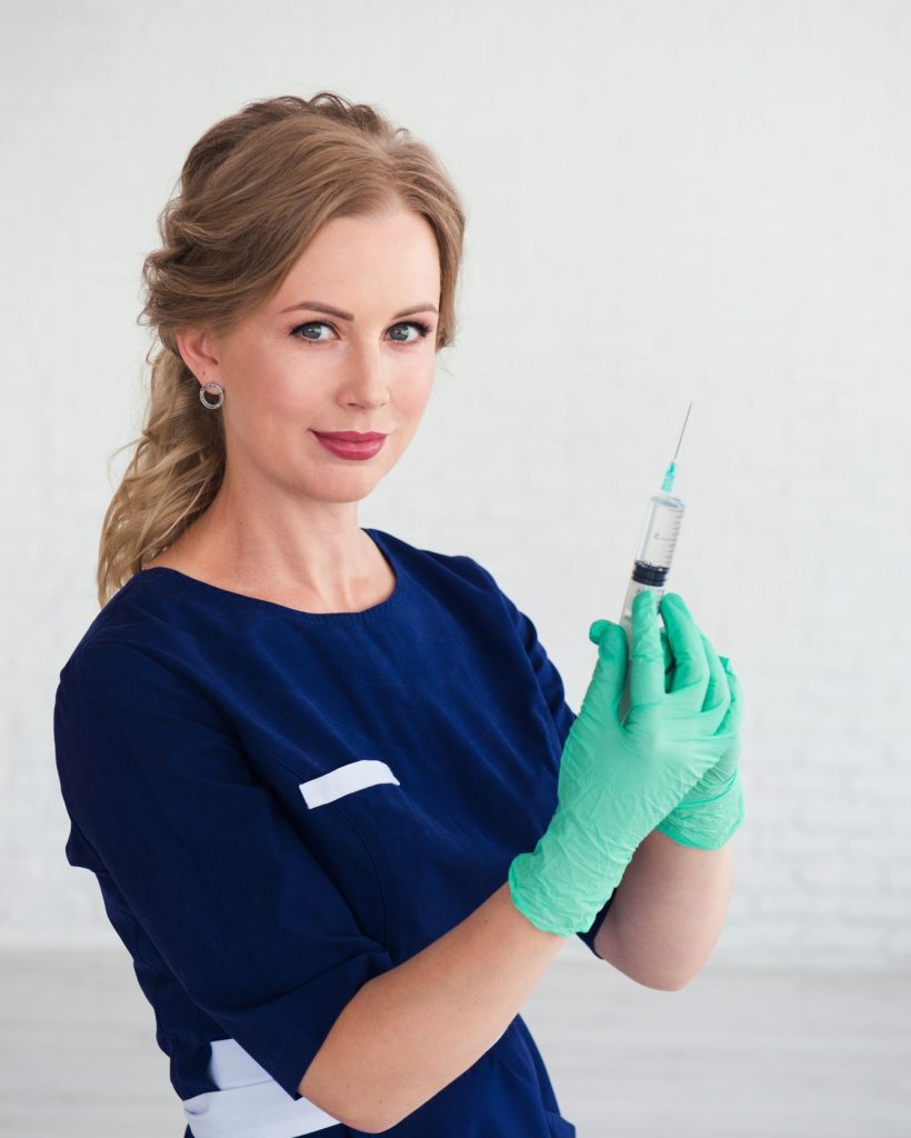 Young woman cosmetologist in blue uniform holding syringe, beauty face injection