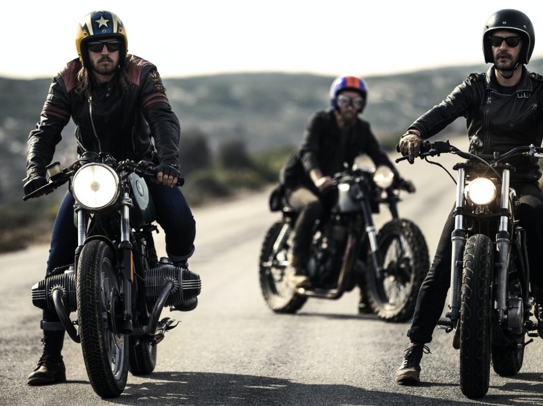Three men wearing open face crash helmets and goggles sitting on cafe racer motorcycles on a rural