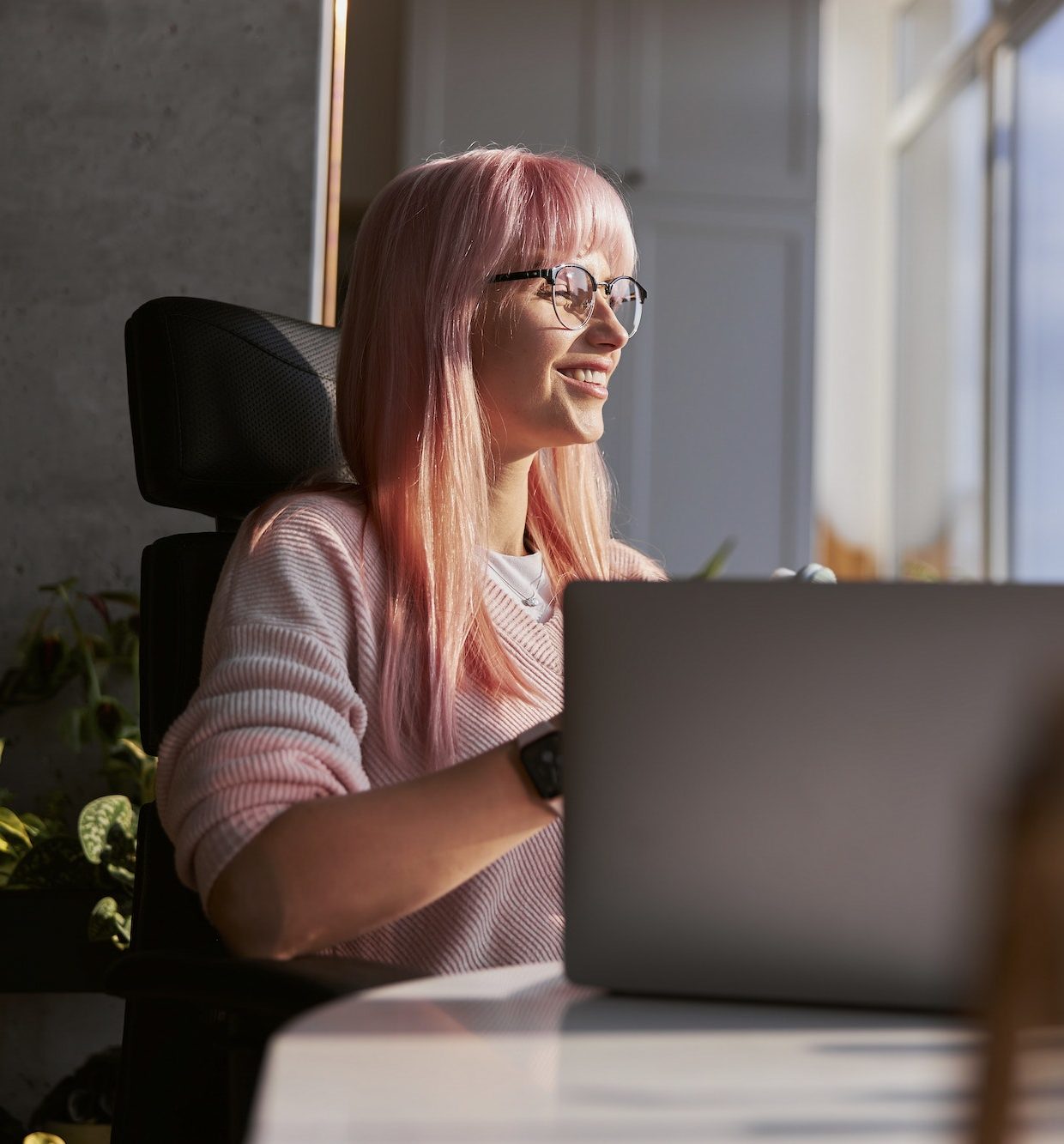 Pink haired woman with elegant glasses works on laptop near window at home