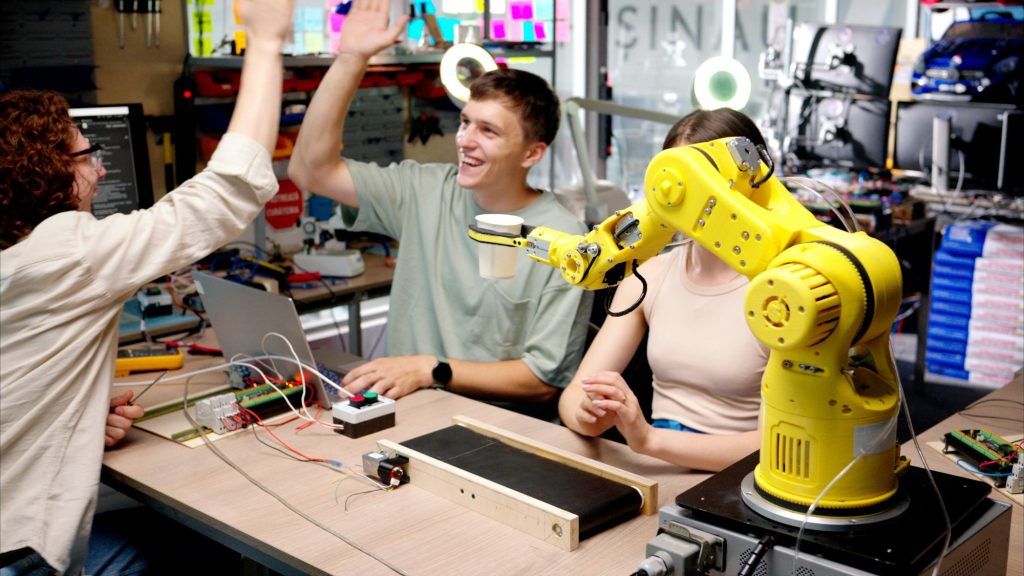 Three young happy engineers fixing an yellow robotic arm in the workshop, computer programming