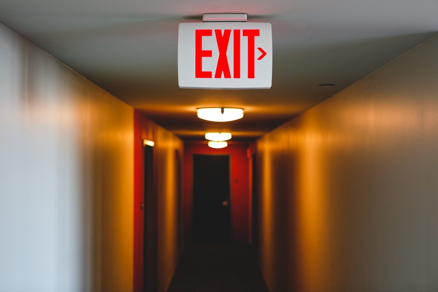 Exit sign in a lit up long hallway, exit to right from eerie hallway