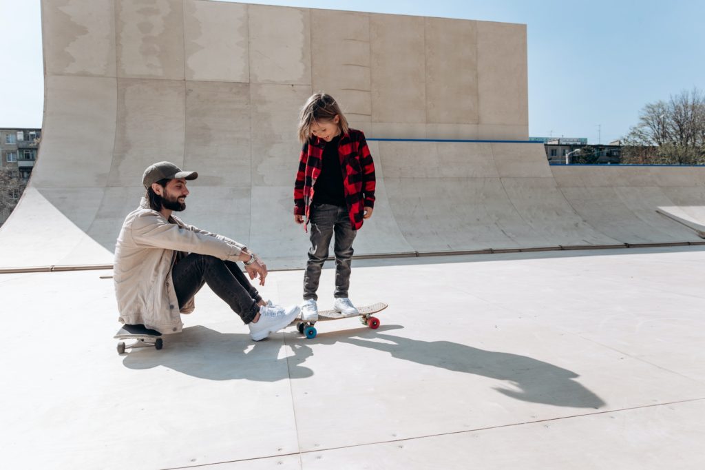 Young father sits on the skateboard and his little son stands on his skateboard in a skate park with
