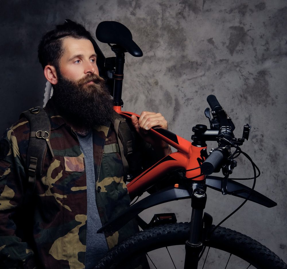 Handsome bearded traveler, dressed in a trendy camouflage jacket, standing with sports bicycle