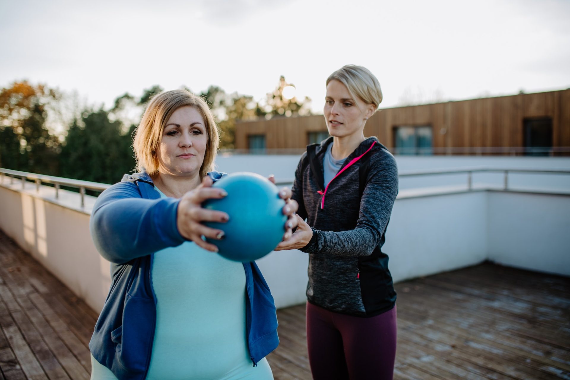 Overweight woman holding ball and exercising with personal trainer in outdoors on gym terrace