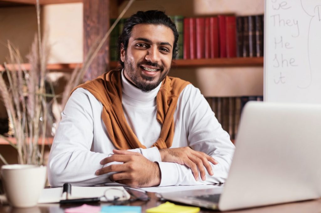 Successful Indian Teacher Sitting At Laptop Posing In Library Indoors