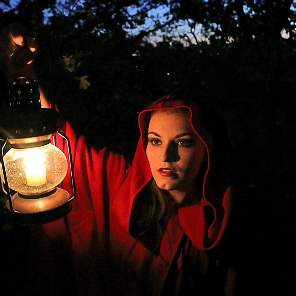 Fairytale portrait of woman in red hood holding lighted lantern as she enters dark woods.