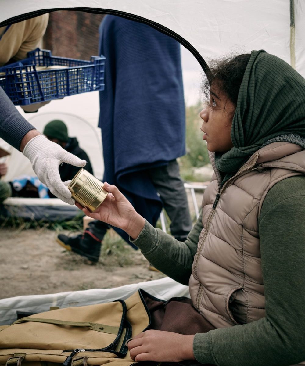 Hungry girl refugee taking tinned food from gloved hand of volunteer
